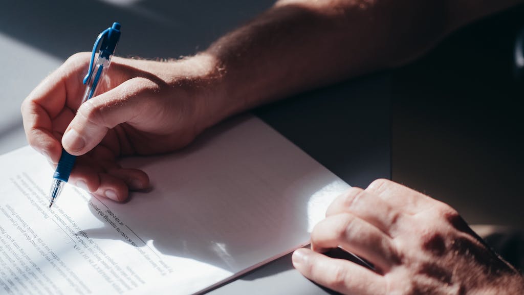 A close-up image of a person's hand signing a document with a blue pen in natural lighting.