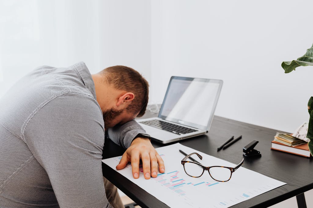A man in a gray shirt sits tiredly at a desk with a laptop and documents, showing signs of workplace stress.